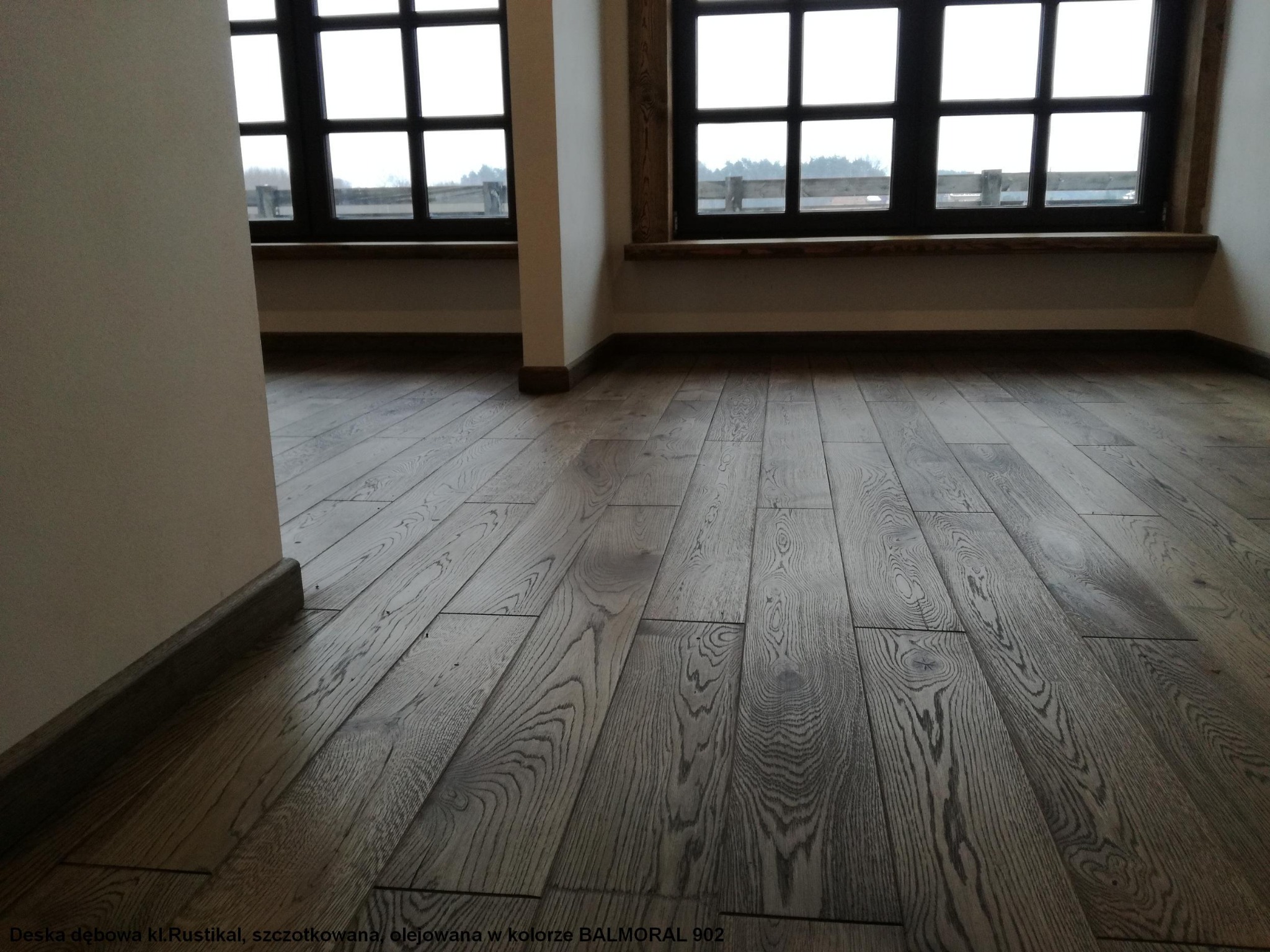 Deska dębowa w klasie Rustikal, szczotkowana i olejowana w kolorze BALMORAL 902, ułożona w jasnym wnętrzu z oknami w ciemnych ramach.