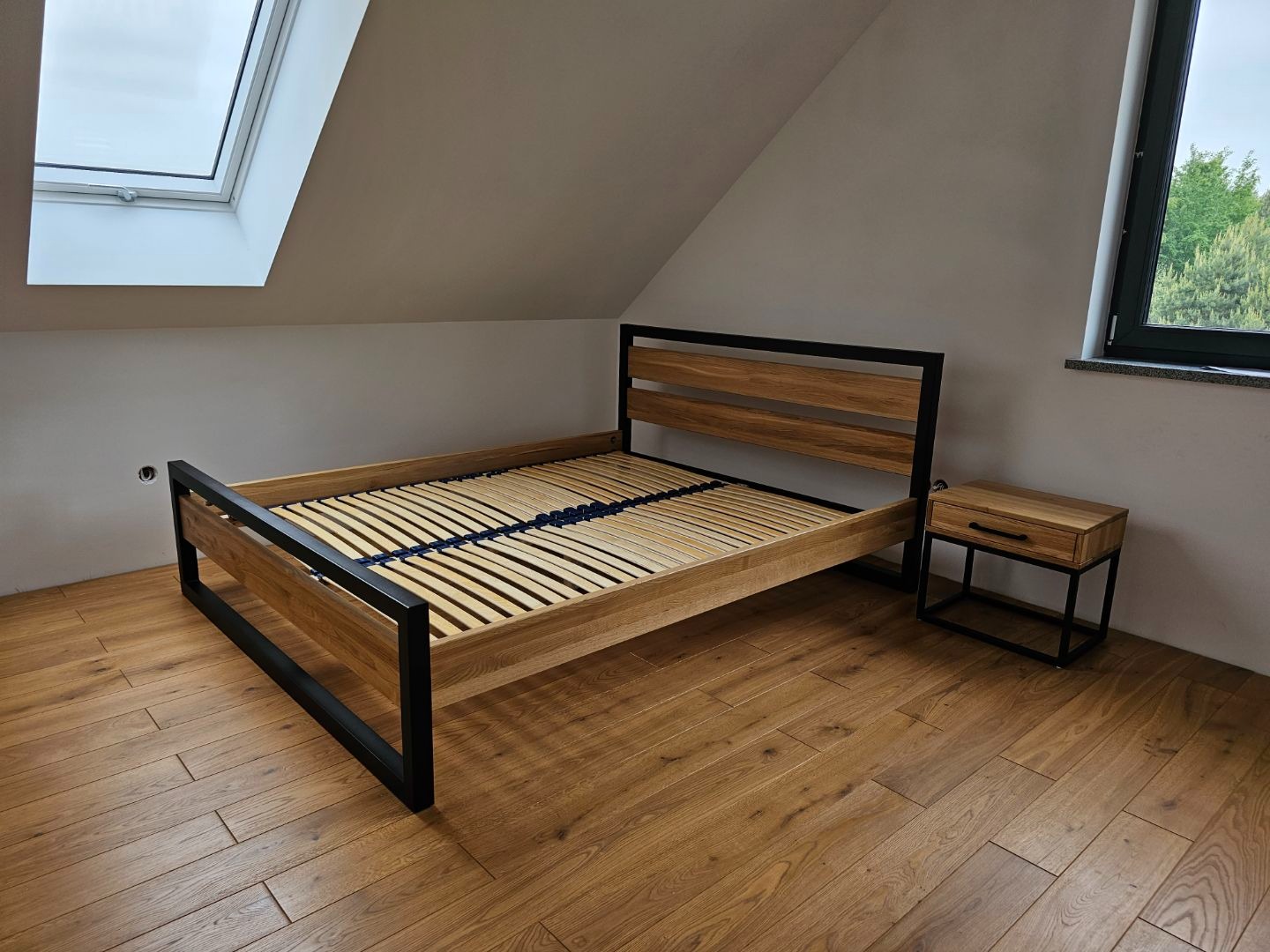 Drewniane łóżko z czarną metalową ramą i dopasowaną szafką nocną na drewnianej podłodze w pokoju na poddaszu z oknami dachowymi.
