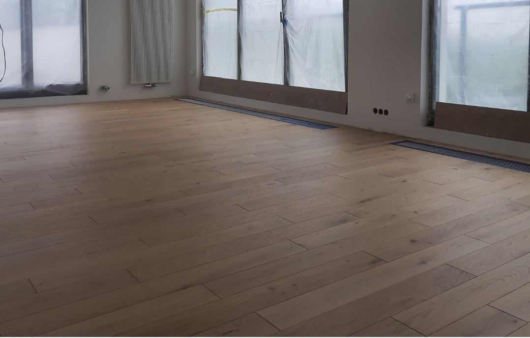 Jasna, drewniana podłoga ułożona w jodełkę w nowym wnętrzu, z widocznymi kratkami wentylacyjnymi w podłodze i oknami zabezpieczonymi folią malarską.