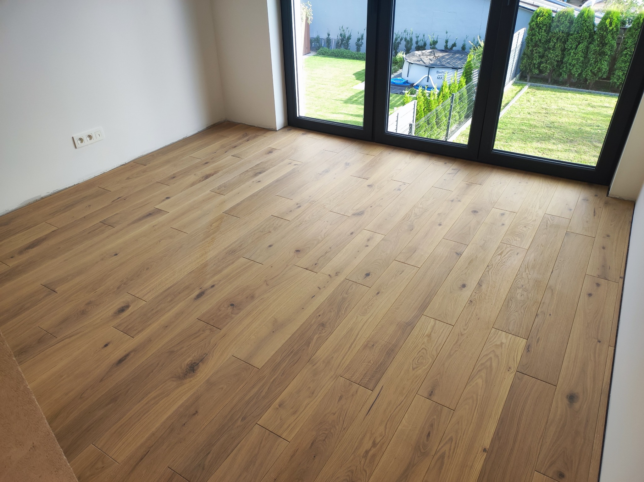 Ułożona drewniana podłoga w jasnym odcieniu dębu, widok z góry, z oknem balkonowym i widokiem na zielony trawnik w tle.