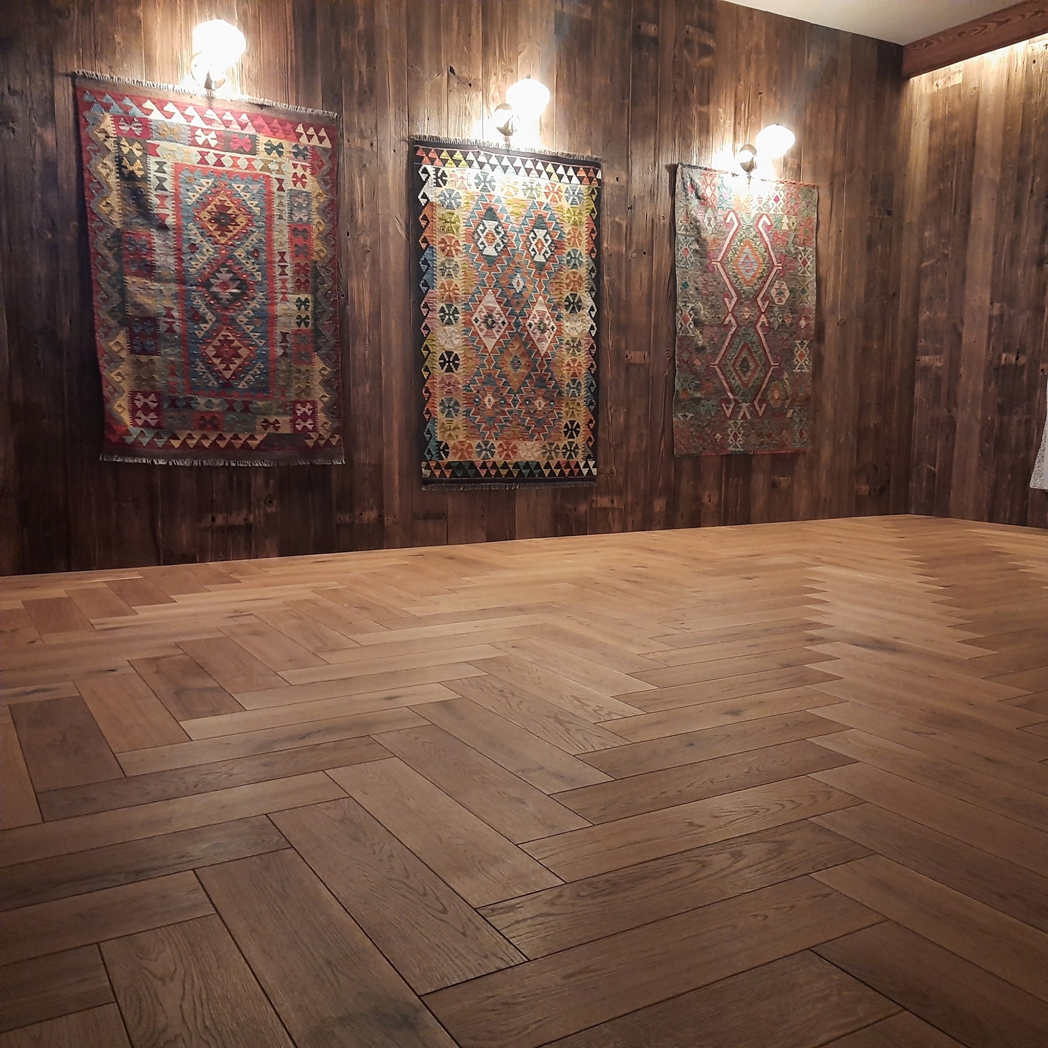 Parkiet ułożony we wzór jodełki francuskiej na tle drewnianej ściany ozdobionej trzema orientalnymi kilimami podświetlonymi kinkietami.