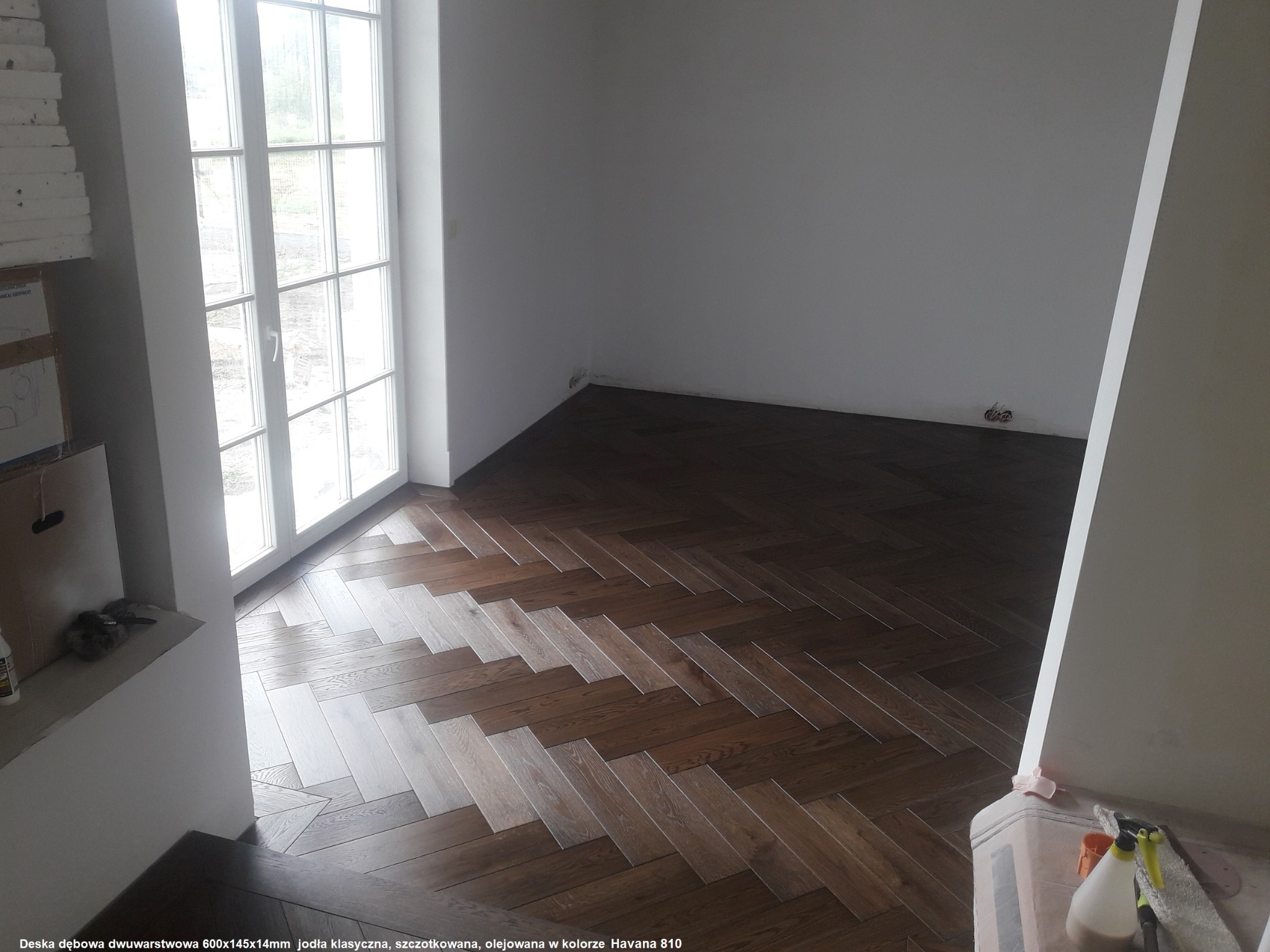 Ułożona jodełka klasyczna z deski dębowej dwuwarstwowej w odcieniu Havana 810, widok z narożnika pomieszczenia z dużym oknem balkonowym.