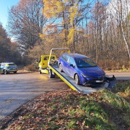 Hol24 Zenon Surmacz - Żółta laweta z uszkodzonym, niebieskim kombi Toyota na platformie, na poboczu drogi, w tle radiowóz policyjny i jesienny krajobraz.