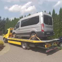 Żółta laweta z srebrnym busem zabezpieczonym pasami transportowymi na platformie, widok z tyłu, w tle zielone tuje i błękitne niebo z chmurami.