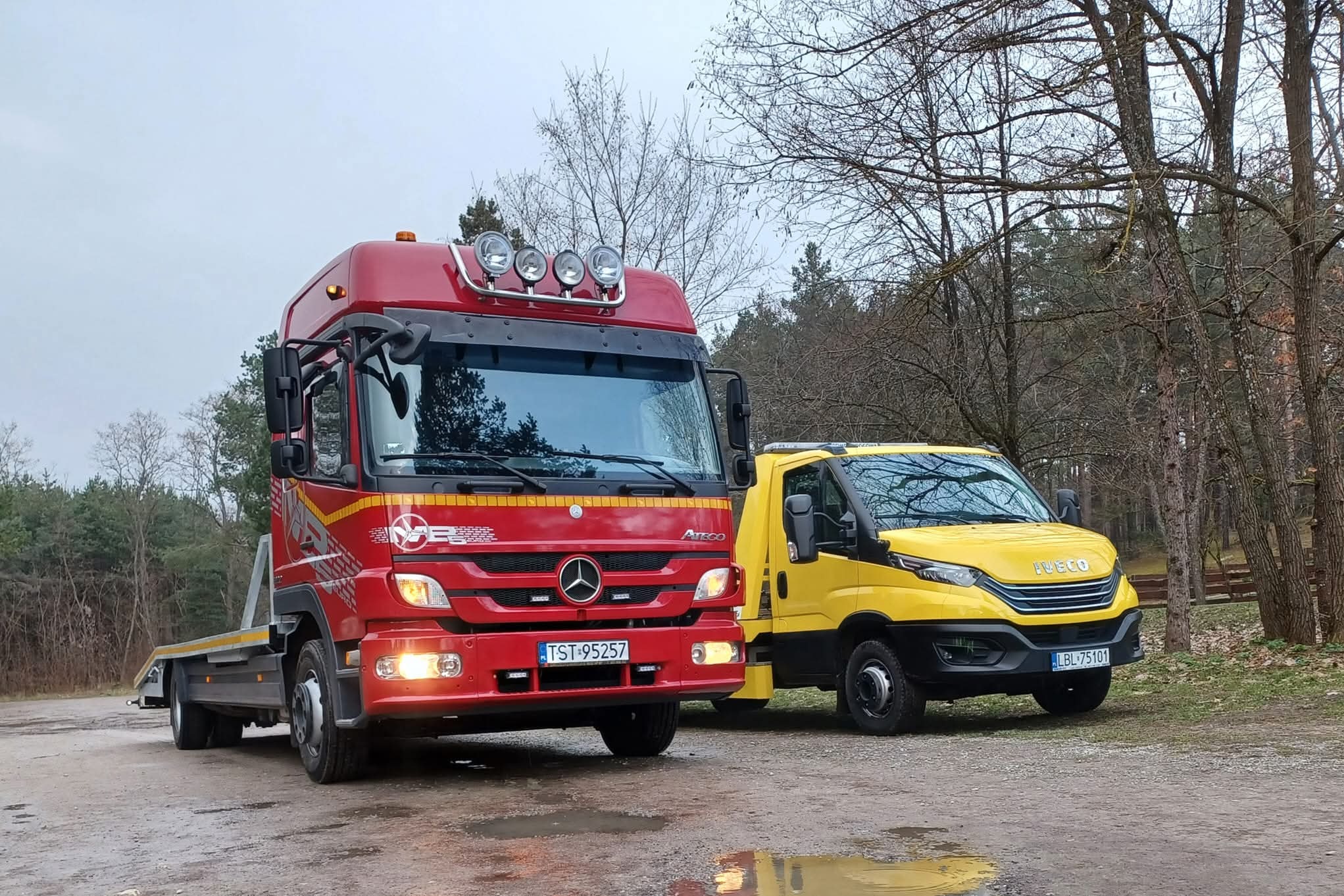 Czerwona laweta Mercedes Atego z czterema reflektorami na dachu, obok żółty bus Iveco na tle drzew i szarego nieba. Tablice rejestracyjne widoczne.