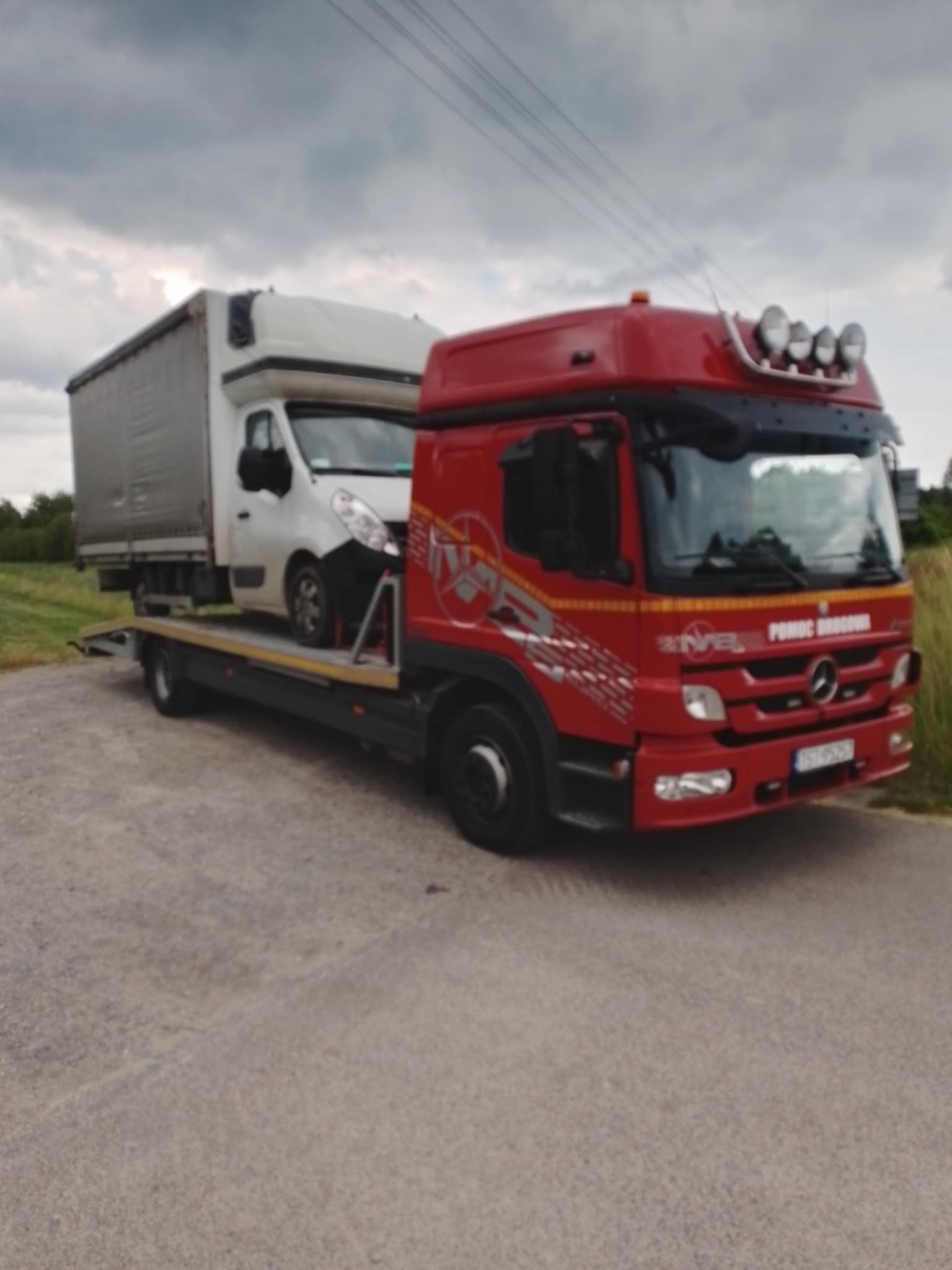 Czerwona laweta Mercedes z białym busem na platformie, zaparkowana na szarej drodze, w tle zielone pole i pochmurne niebo. Widoczne światła na dachu lawety i grafika na kabinie.