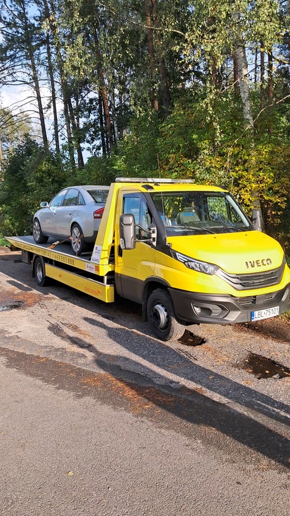 Żółta laweta Iveco z srebrnym samochodem osobowym na platformie, zaparkowana na poboczu drogi w słoneczny dzień, z drzewami w tle. Widoczny numer rejestracyjny.