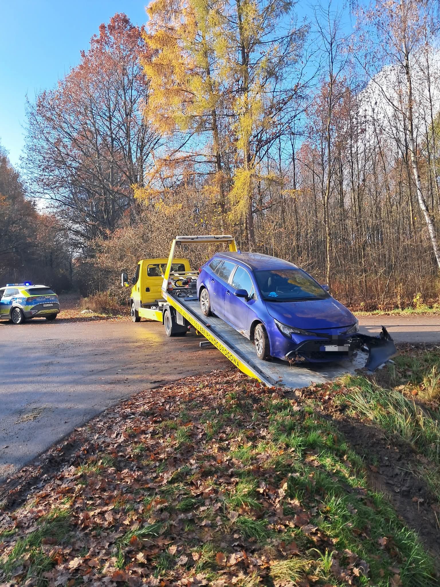 Żółta laweta z uszkodzonym, niebieskim kombi Toyota na platformie, na poboczu drogi, w tle radiowóz policyjny i jesienny krajobraz.