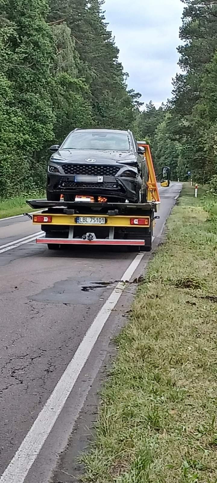 Czarny uszkodzony samochód Hyundai Kona na żółtej lawecie z numerem rejestracyjnym LBL 75101, sfotografowany na poboczu drogi w otoczeniu drzew.