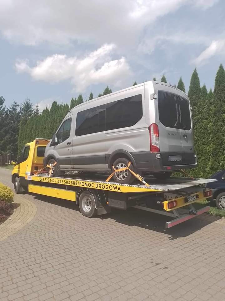 Żółta laweta z srebrnym busem zabezpieczonym pasami transportowymi na platformie, widok z tyłu, w tle zielone tuje i błękitne niebo z chmurami.