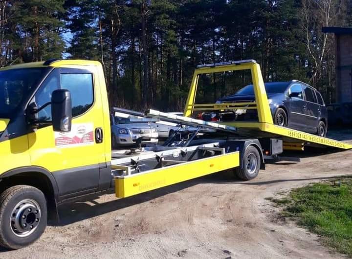 Żółta laweta z załadowanym samochodem osobowym na polnej drodze, częściowo widoczny drugi pojazd na platformie.