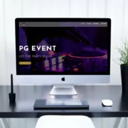 Ekran komputera iMac wyświetlający stronę internetową pg-event.pl z motywem imprezowym, na czarnym biurku z lampką, tabletem graficznym, myszką, kwiatami w wazonie i rośliną w doniczce.