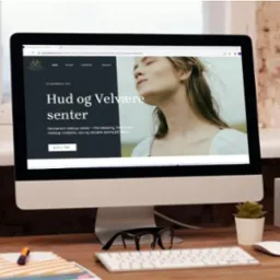 Monitor wyświetlający stronę internetową hudogvelvaresenter.no z motywem beauty i wizerunkiem kobiety, stojący na biurku z klawiaturą, myszką, okularami i dekoracjami.