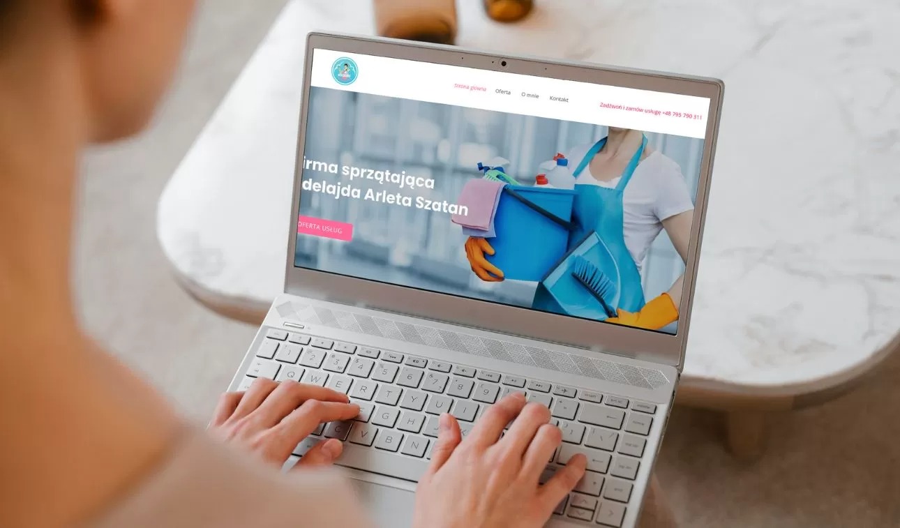Osoba z tyłu głowy przegląda na laptopie stronę internetową firmy sprzątającej adelajda-sprzatanie.pl, na ekranie widoczna kobieta w fartuchu trzymająca wiadro ze środkami czystości.