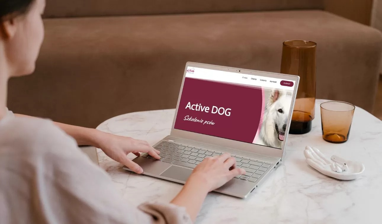 Kobieta przegląda na laptopie stronę internetową active-dog.pl z wizerunkiem białego owczarka szwajcarskiego, obok marmurowy stolik z dekoracyjnym wazonem i ceramiczną dłonią trzymającą słuchawki.