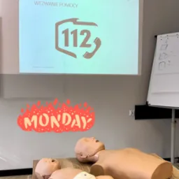 Sala szkoleniowa z manekinami niemowlęcia i dorosłego do nauki pierwszej pomocy, rzutnik wyświetla numer alarmowy 112, obok apteczka pierwszej pomocy.