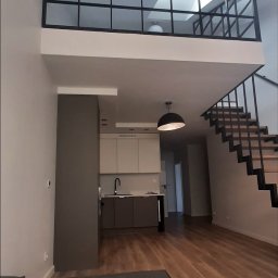 Krzysztof Kowalski - Dwupoziomowe wnętrze mieszkania z antresolą oddzieloną czarną, przeszkloną konstrukcją. Widoczne schody z czarną balustradą, kuchnia i drewniana podłoga.