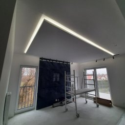 Krzysztof Kowalski - Nowoczesne wykończenie wnętrza: podwieszany sufit z oświetleniem LED, ciemna ściana z oknem, rusztowanie w pokoju. Etap prac wykończeniowych.