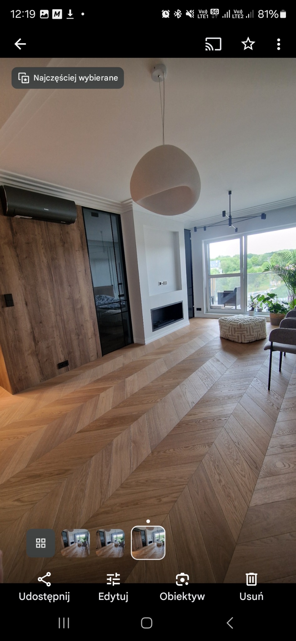 Salon z parkietem ułożonym we wzór jodełki, nowoczesnym oświetleniem i minimalistycznym kominkiem. Widok na balkon z roślinami. Nowoczesne wykończenie wnętrza.