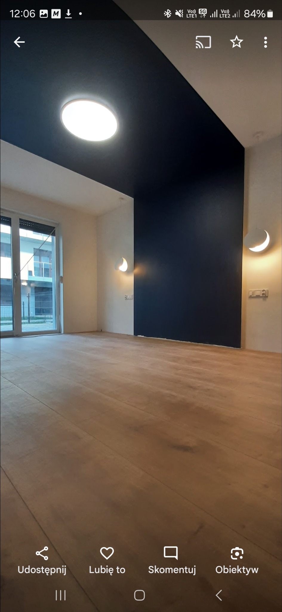 Wykończone wnętrze: parkiet, ciemna ściana i sufit, białe ściany boczne z lampami. Nowoczesny design, jasne światło. Widok na okno balkonowe.