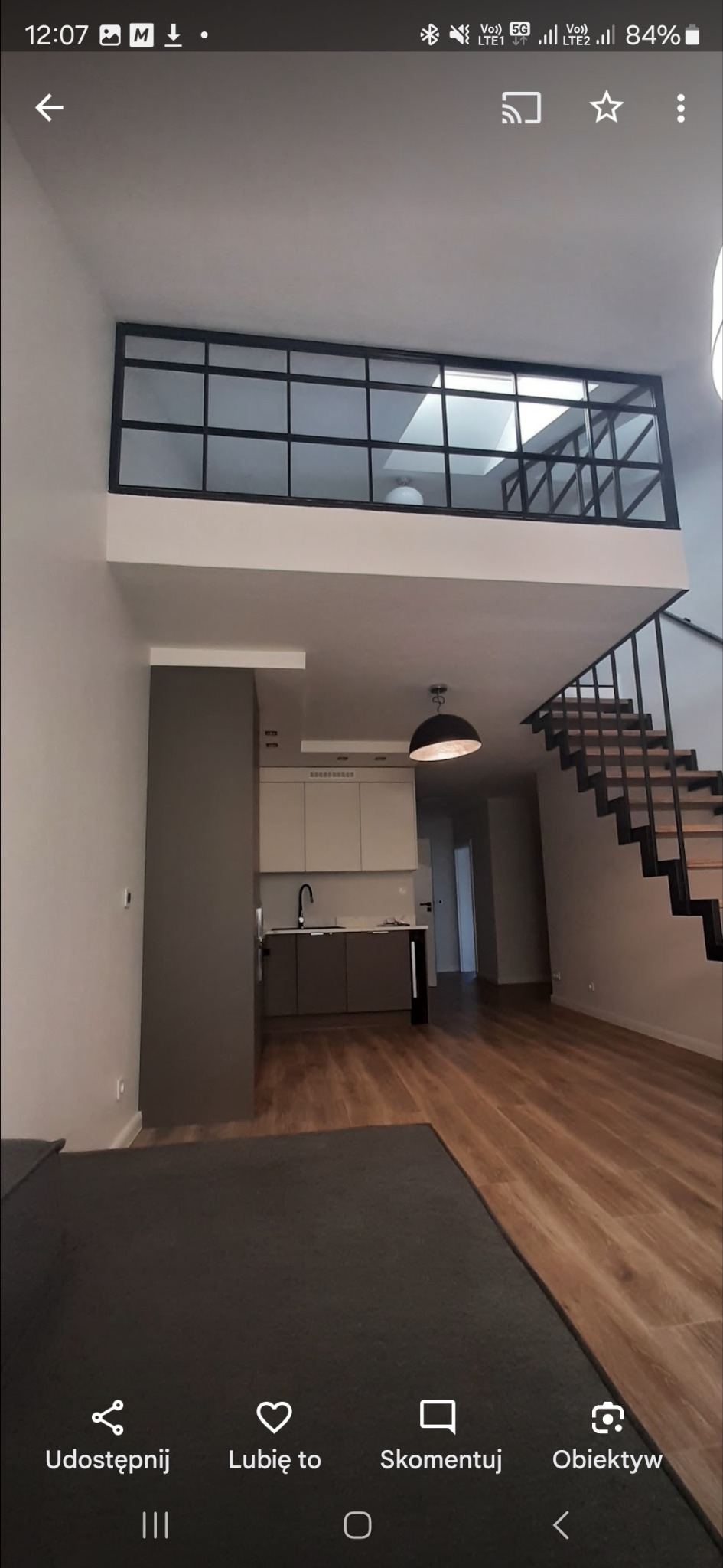 Dwupoziomowe wnętrze mieszkania z antresolą oddzieloną czarną, przeszkloną konstrukcją. Widoczne schody z czarną balustradą, kuchnia i drewniana podłoga.