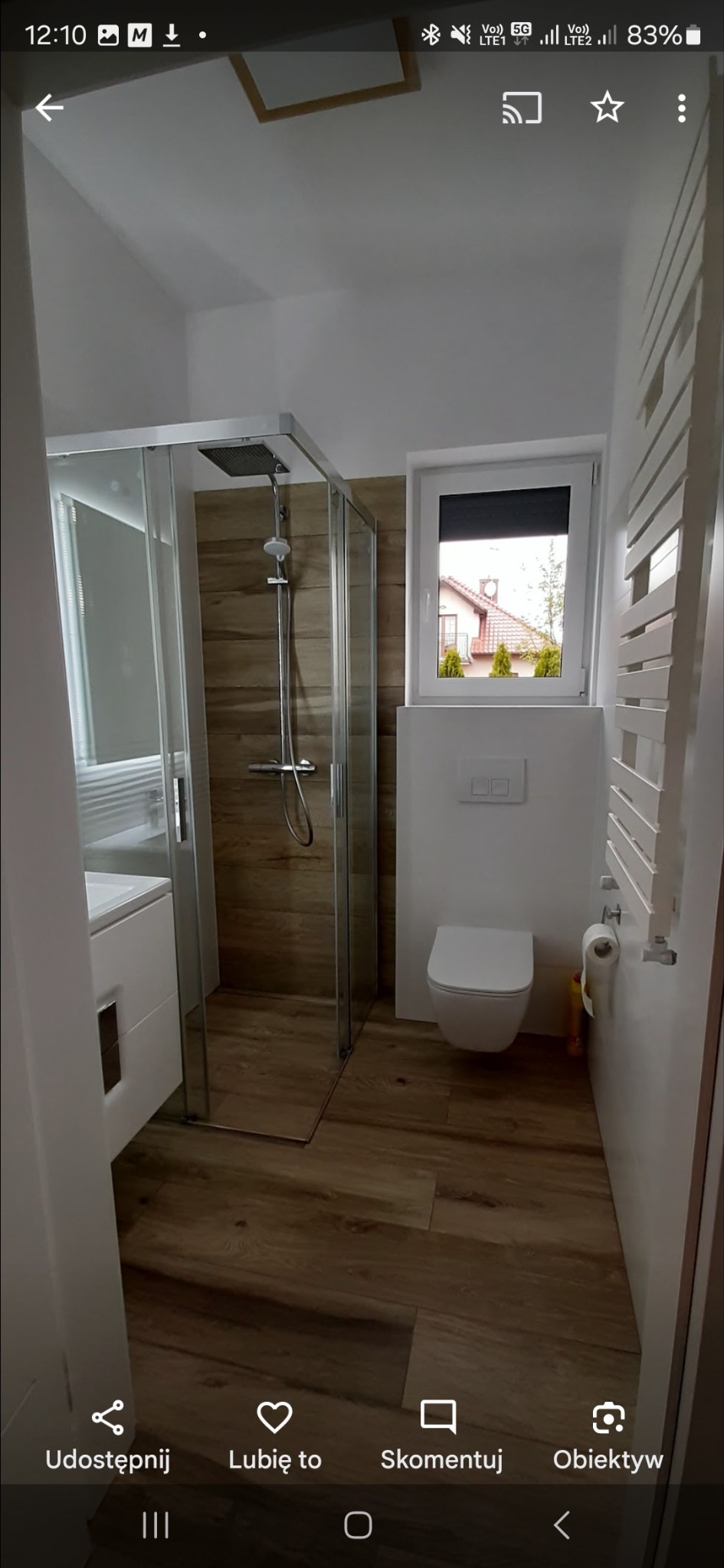 Nowoczesna łazienka z kabiną prysznicową typu walk-in, drewnopodobną podłogą i białymi ścianami. Widoczne okno z roletą i grzejnik drabinkowy.