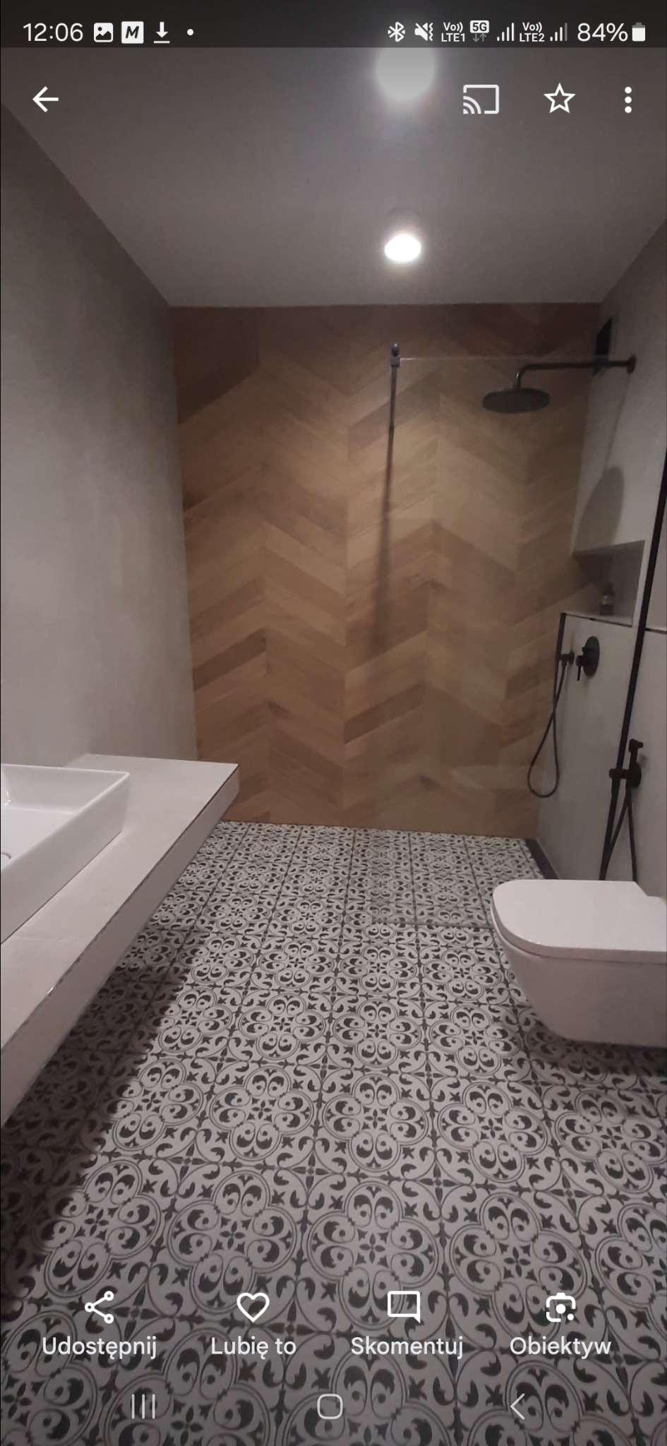 Nowoczesna łazienka po remoncie: drewniana ściana w jodełkę, wzorzysta podłoga, biała umywalka i toaleta. Czarna armatura dodaje elegancji.