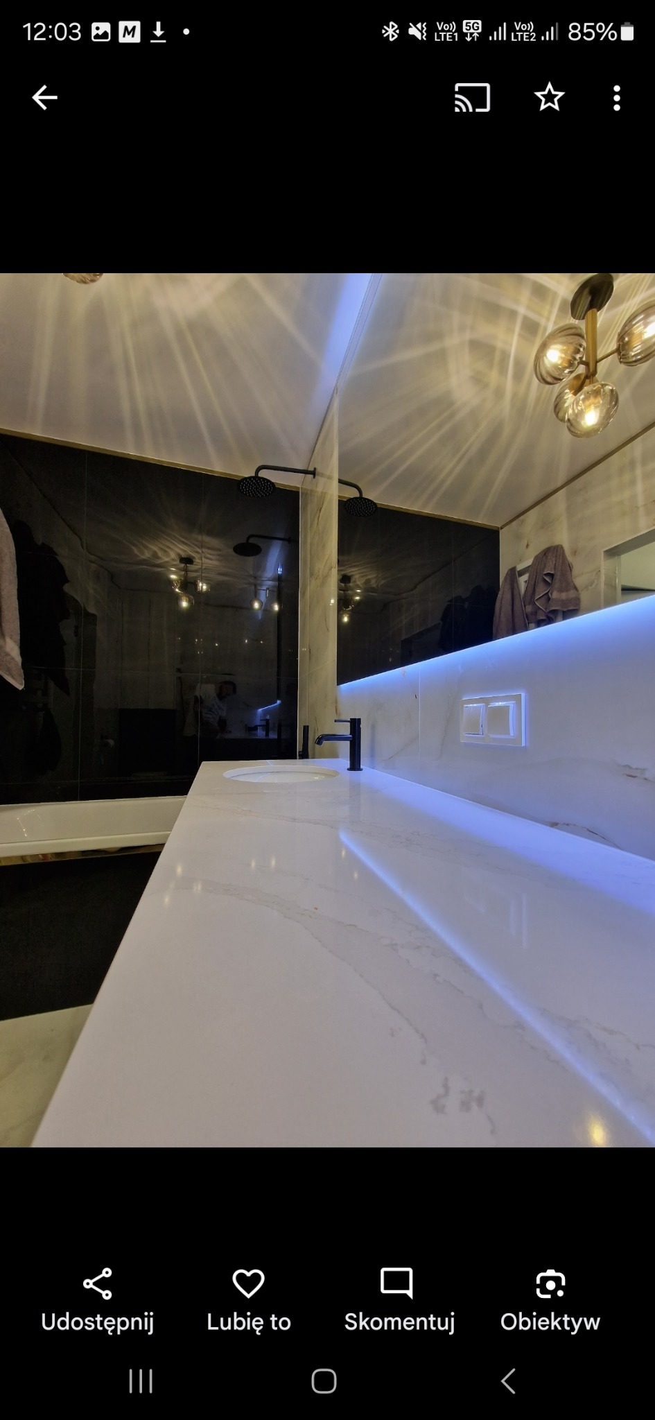 Nowoczesna łazienka z czarnymi płytkami, marmurowym blatem i podświetleniem LED. Widoczna armatura w kolorze czarnym i designerska lampa sufitowa.