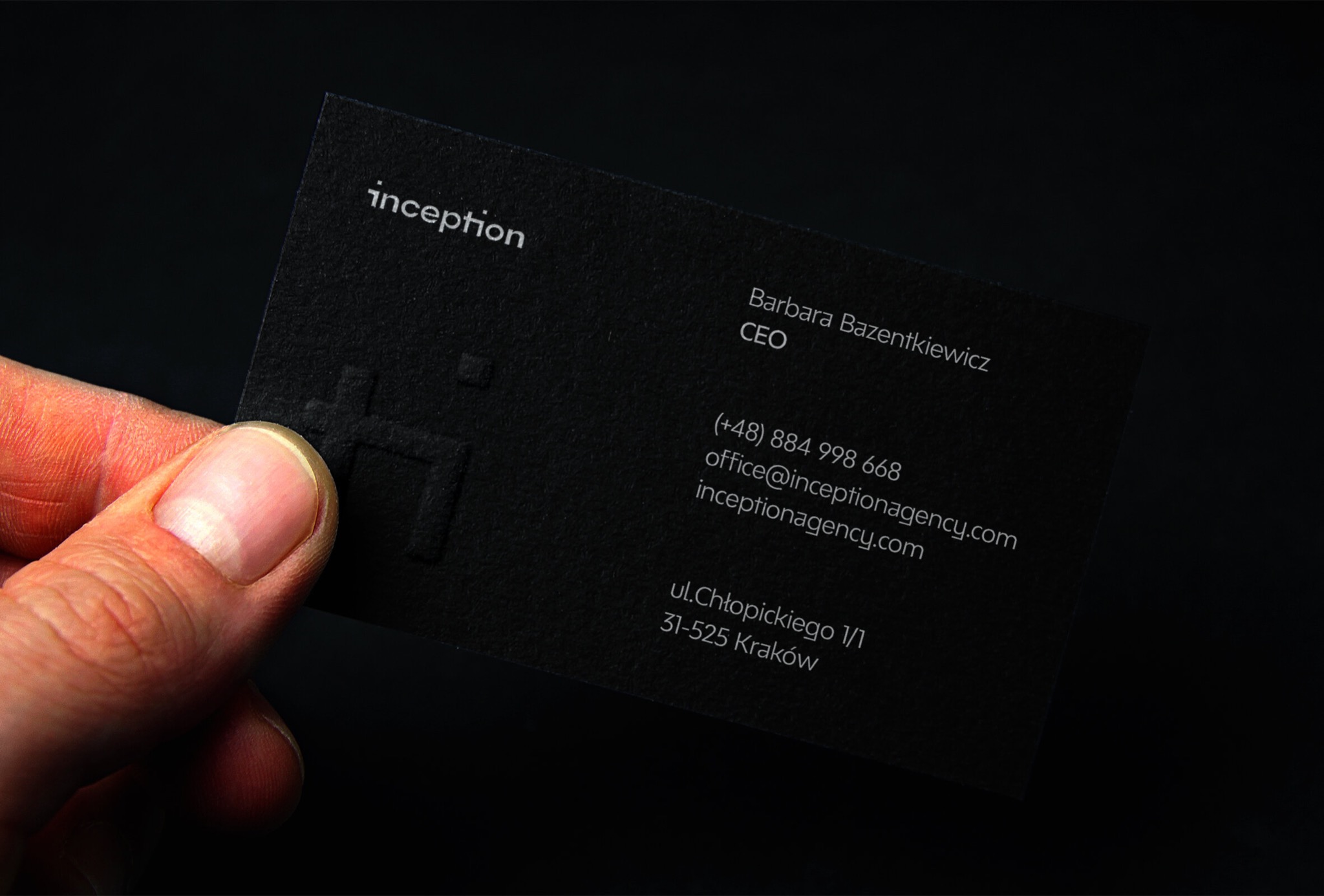 Czarny, matowy wizytownik z tłoczonym logo 'inception', trzymany w palcach, z białym tekstem zawierającym imię, nazwisko, stanowisko, numer telefonu, adres e-mail i adres firmy na ciemnym tle.