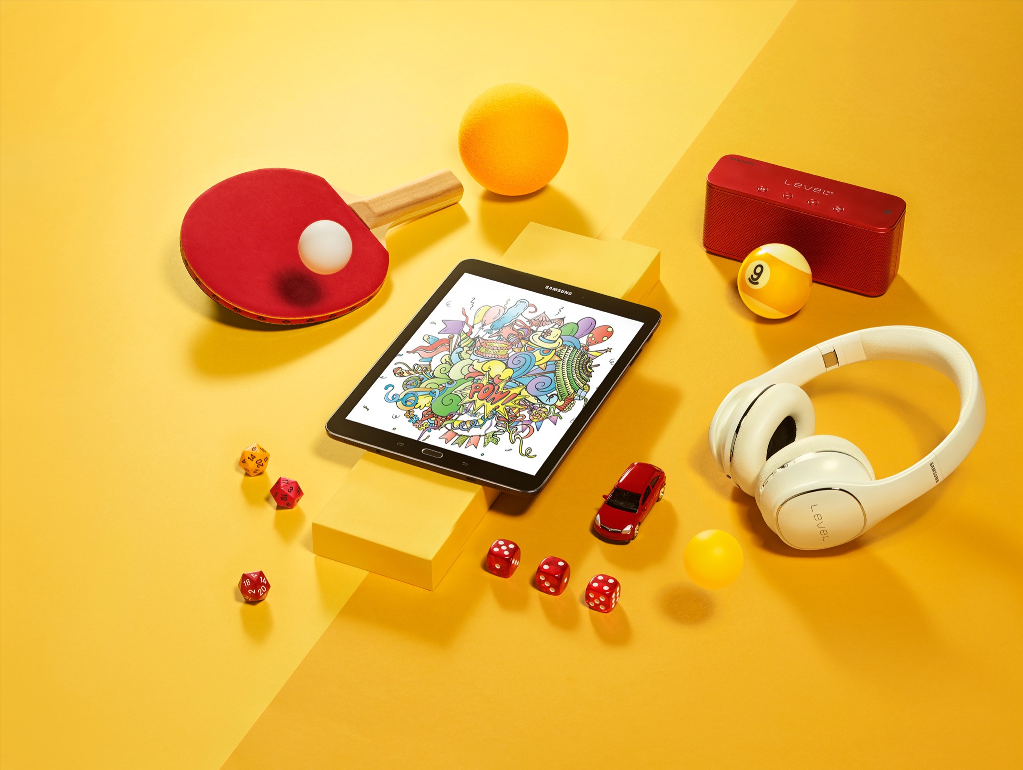 Żółte tło ze starannie rozmieszczonymi przedmiotami: tablet z kolorową grafiką, słuchawki, głośnik, piłeczka bilardowa z numerem 9, rakietka do tenisa stołowego, piłeczki, kostki do gry i model...