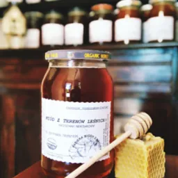 Słoik miodu leśnego z etykietą, drewniany nabierak i plaster wosku pszczelego na tle półek z innymi słoikami miodu.