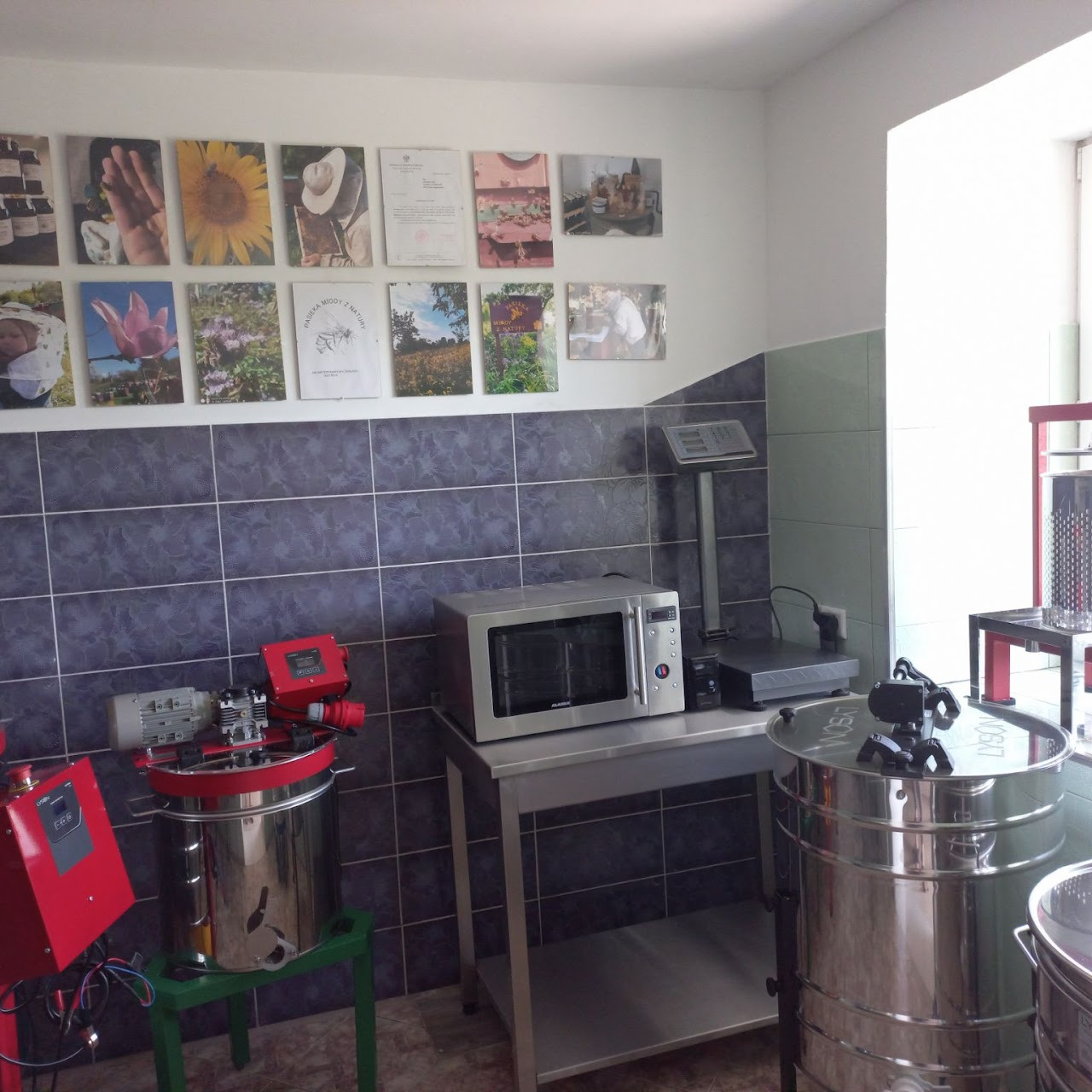 Wnętrze pracowni z wirówkami do miodu, wagą elektroniczną i kuchenką mikrofalową na tle ściany z fotografiami o tematyce pszczelarskiej.