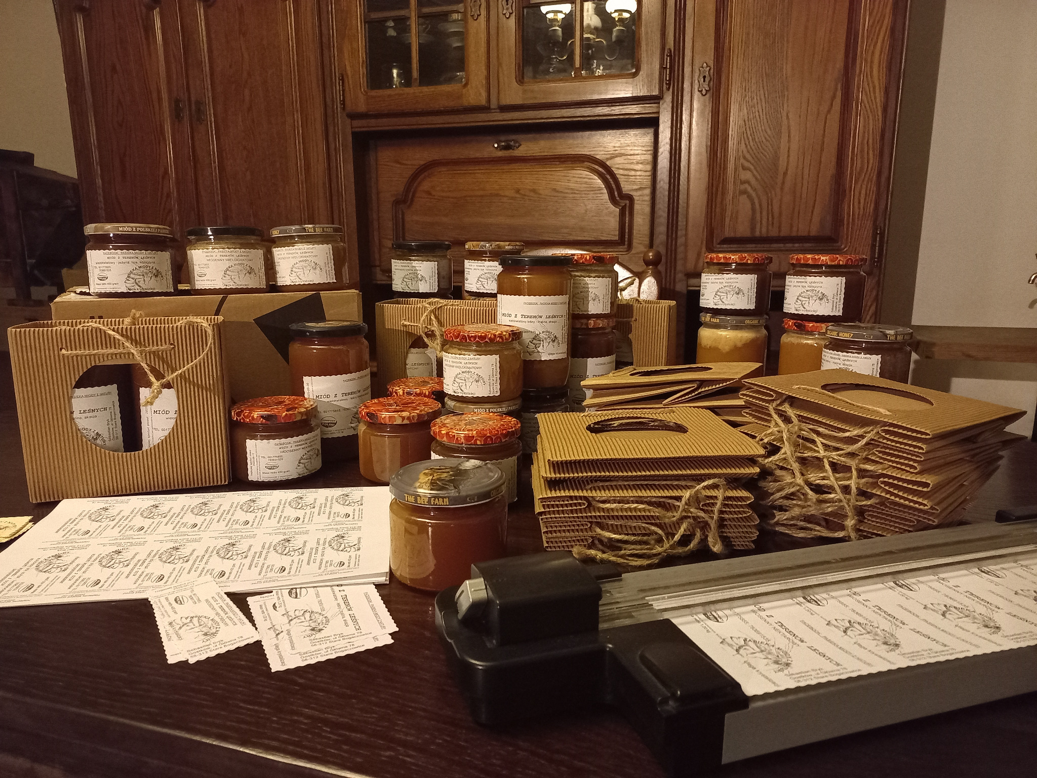 Słoiki z różnymi rodzajami miodu, kartonowe opakowania i etykiety do naklejenia na tle drewnianej szafki, widoczny również obcinak do papieru.