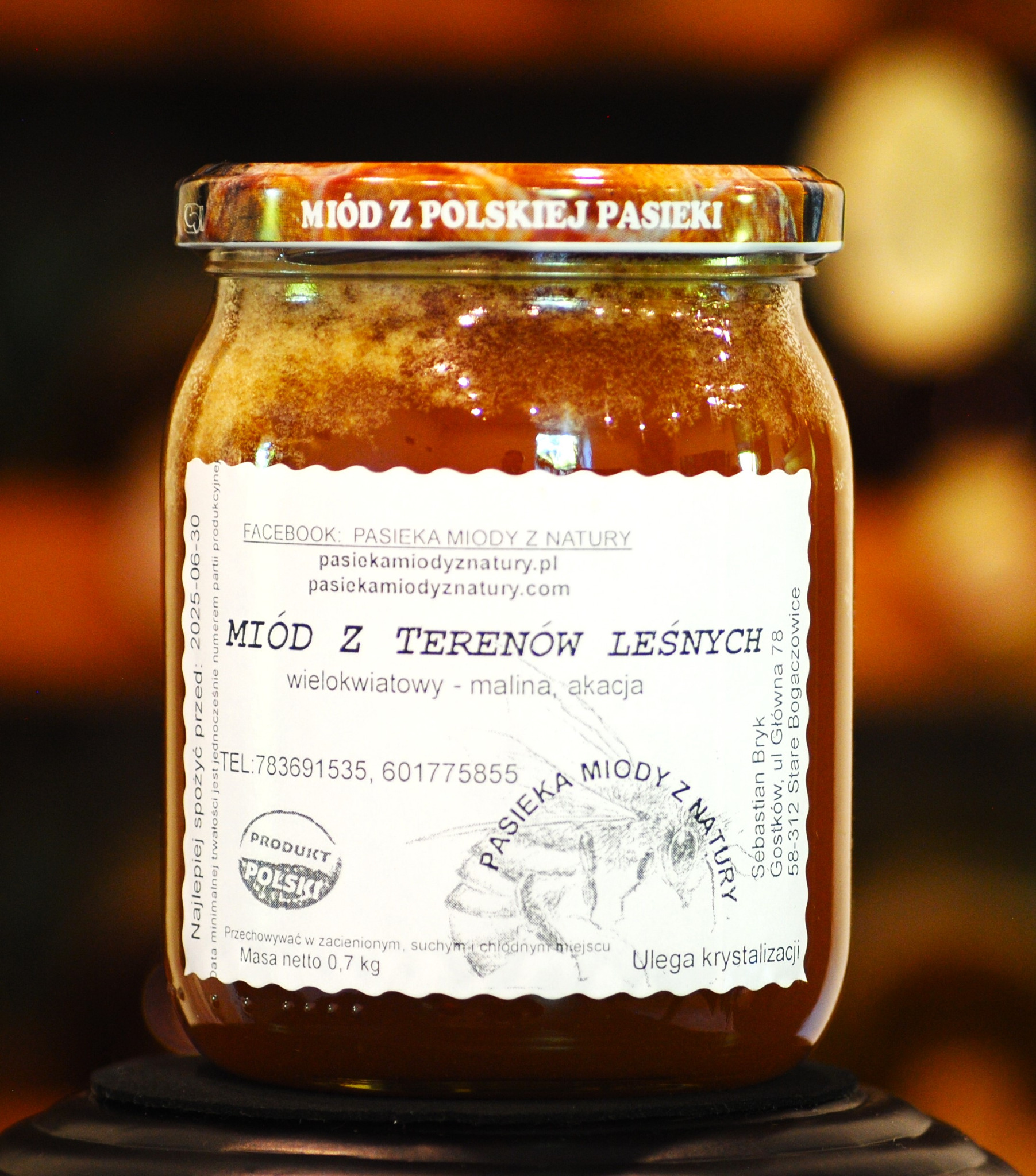 Słoik z ciemnym, gęstym miodem z polskiej pasieki, etykieta informuje o miodzie z terenów leśnych, wielokwiatowym z maliną i akacją, widoczny adres strony internetowej i dane kontaktowe pasieki.