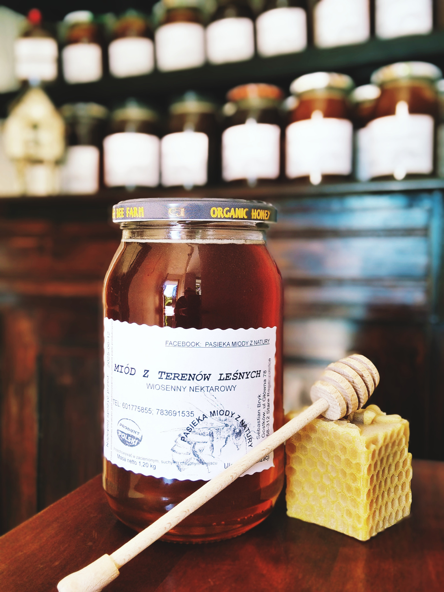 Słoik miodu leśnego z etykietą, drewniany nabierak i plaster wosku pszczelego na tle półek z innymi słoikami miodu.