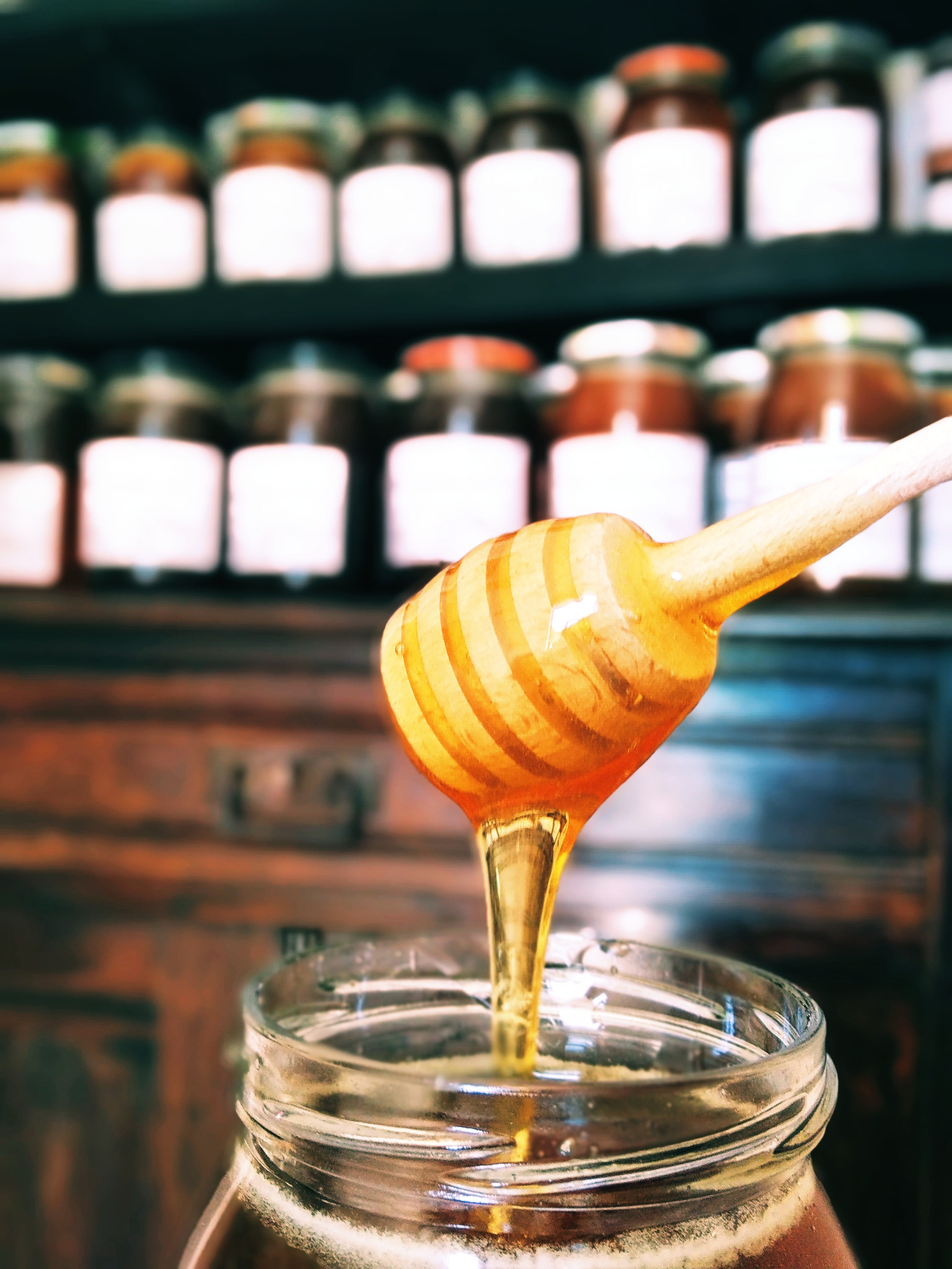 Klarowna strużka cieknącego płynnego złocistego słodkiego produktu spływa z drewnianego nabieraka do słoika, w tle rzędy słoików na ciemnej półce.