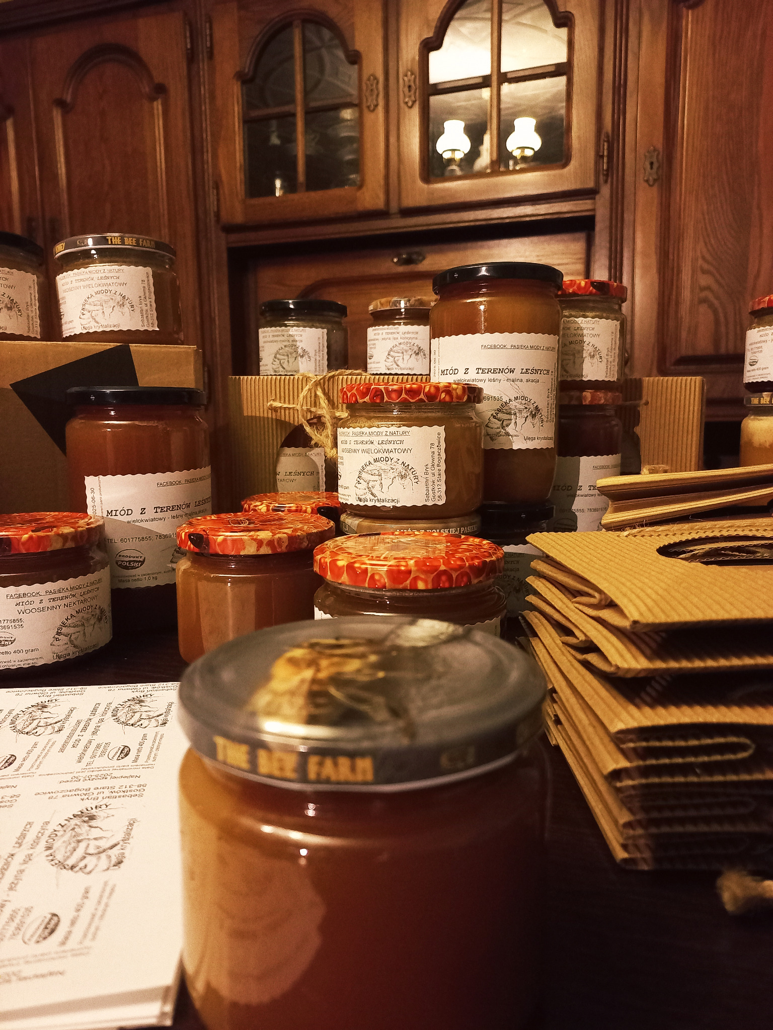 Słoiki z miodem od lokalnego producenta, z etykietami informującymi o rodzaju miodu i składzie, ustawione na drewnianym blacie na tle rustykalnej szafki z oświetleniem.