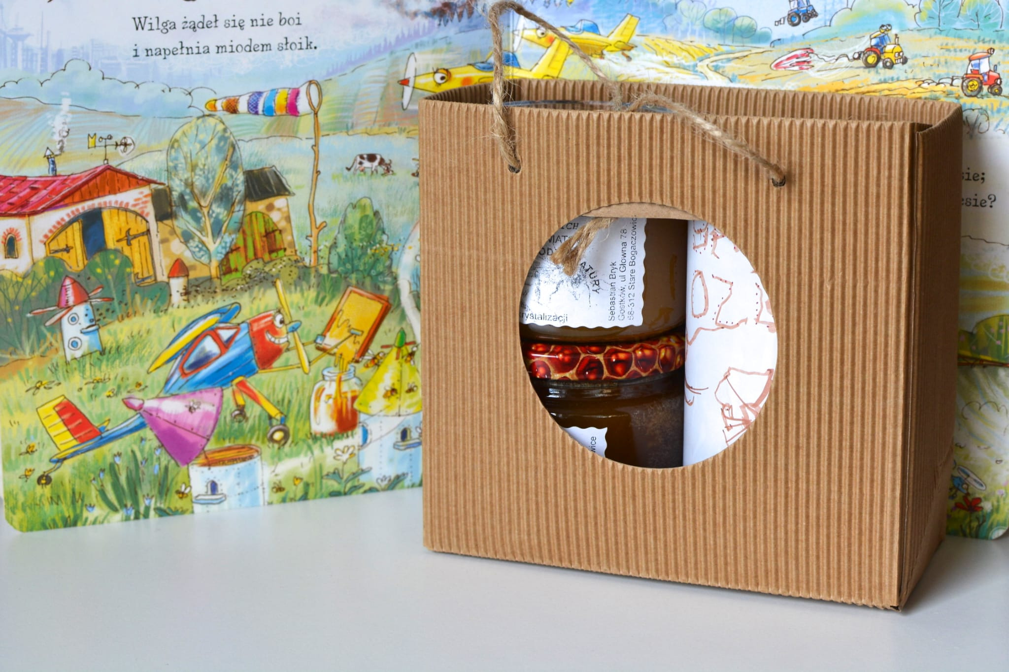 Słoiki z miodem w tekturowej torbie z okienkiem, na tle otwartej książki z ilustracjami wiejskiego krajobrazu i samolotów