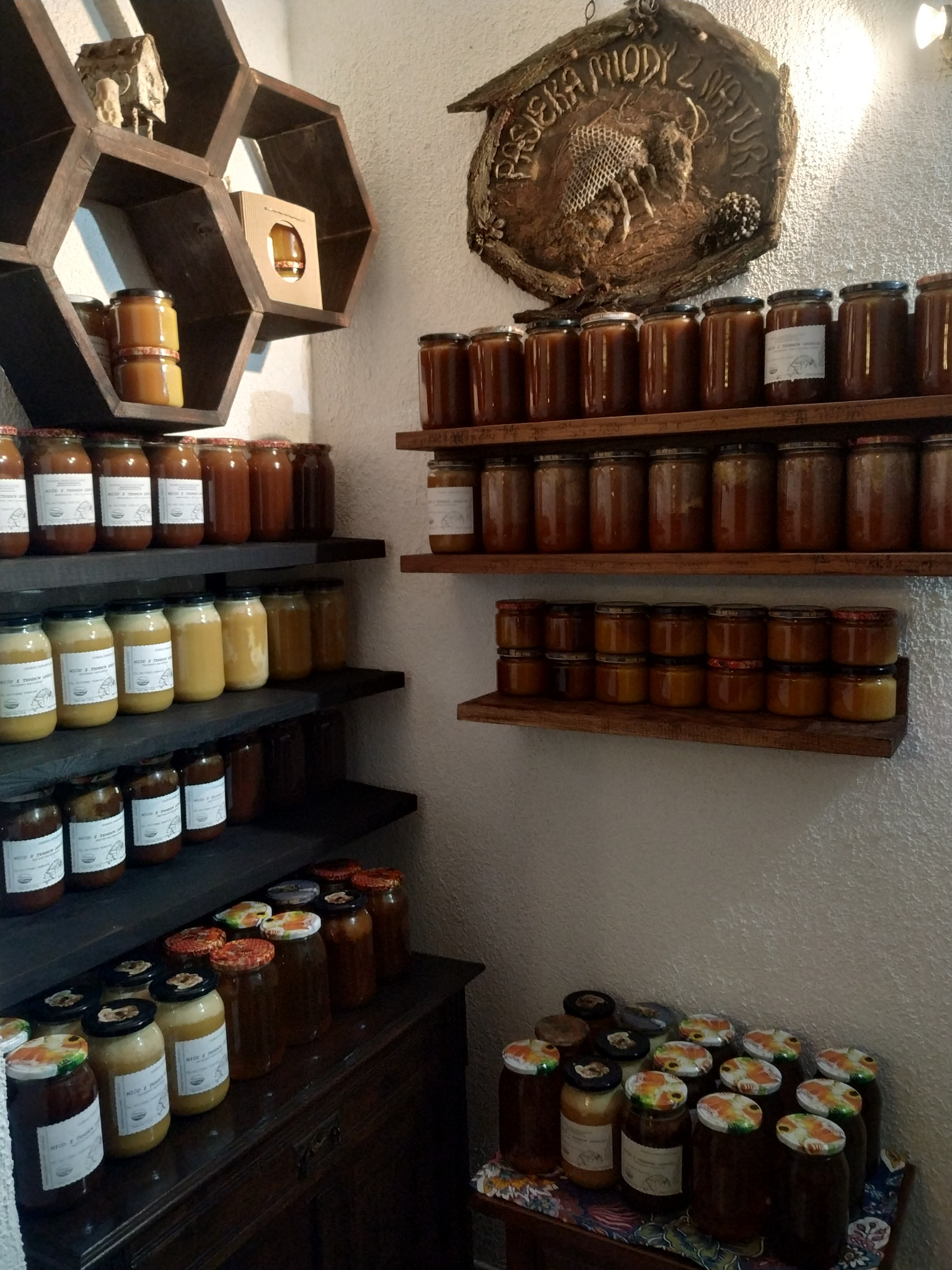 Słoiki z miodem różnych gatunków na drewnianych półkach i komodzie, dekoracyjna tablica z napisem 'Pasieka Miodu z Natury' w tle.