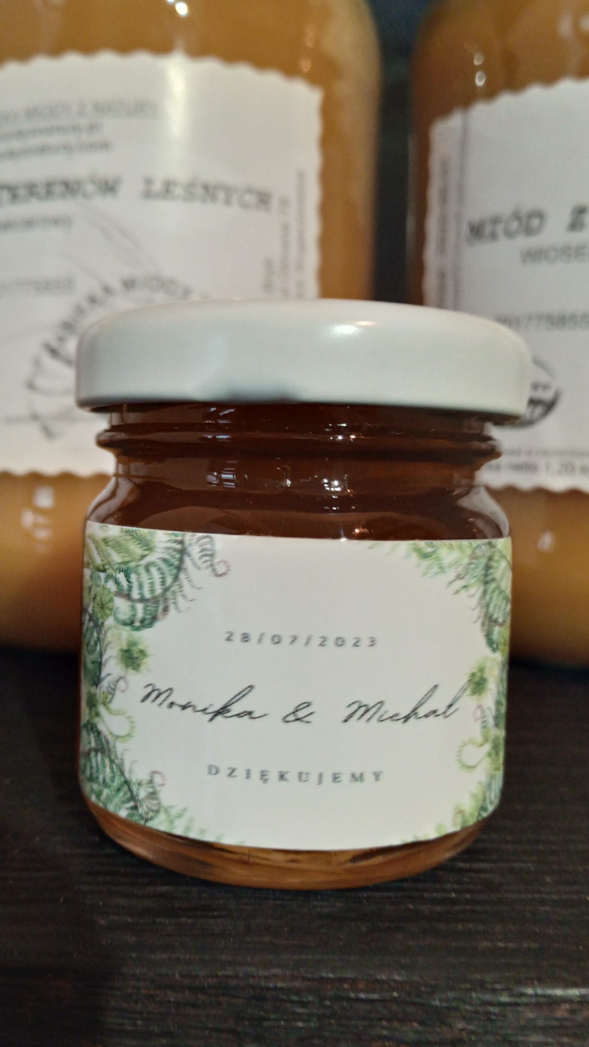 Słoik z ciemnym miodem, etykieta z paprociami, datą 28.07.2023 oraz imionami Monika i Michał, podziękowanie, w tle większe słoiki z jasnym miodem.