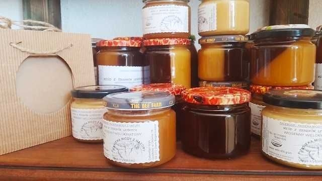 Różne słoiki z miodem o różnych odcieniach, z etykietami i wzorzystymi pokrywkami, ustawione na drewnianej półce, obok papierowej torby z wyciętym okienkiem.