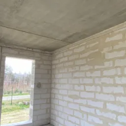 Surowy stan wnętrza: ściany z białych bloczków, betonowy sufit z widocznymi przewodami elektrycznymi w peszlach, otwór okienny z widokiem na zewnątrz.