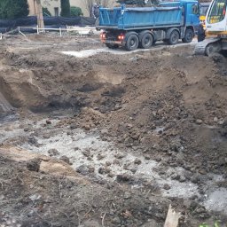 K.S Nadzór budowlany - Wykop fundamentowy z widocznymi śladami po pracy koparki, w tle ciężarówka wywożąca urobek, na placu budowy w pochmurny dzień.