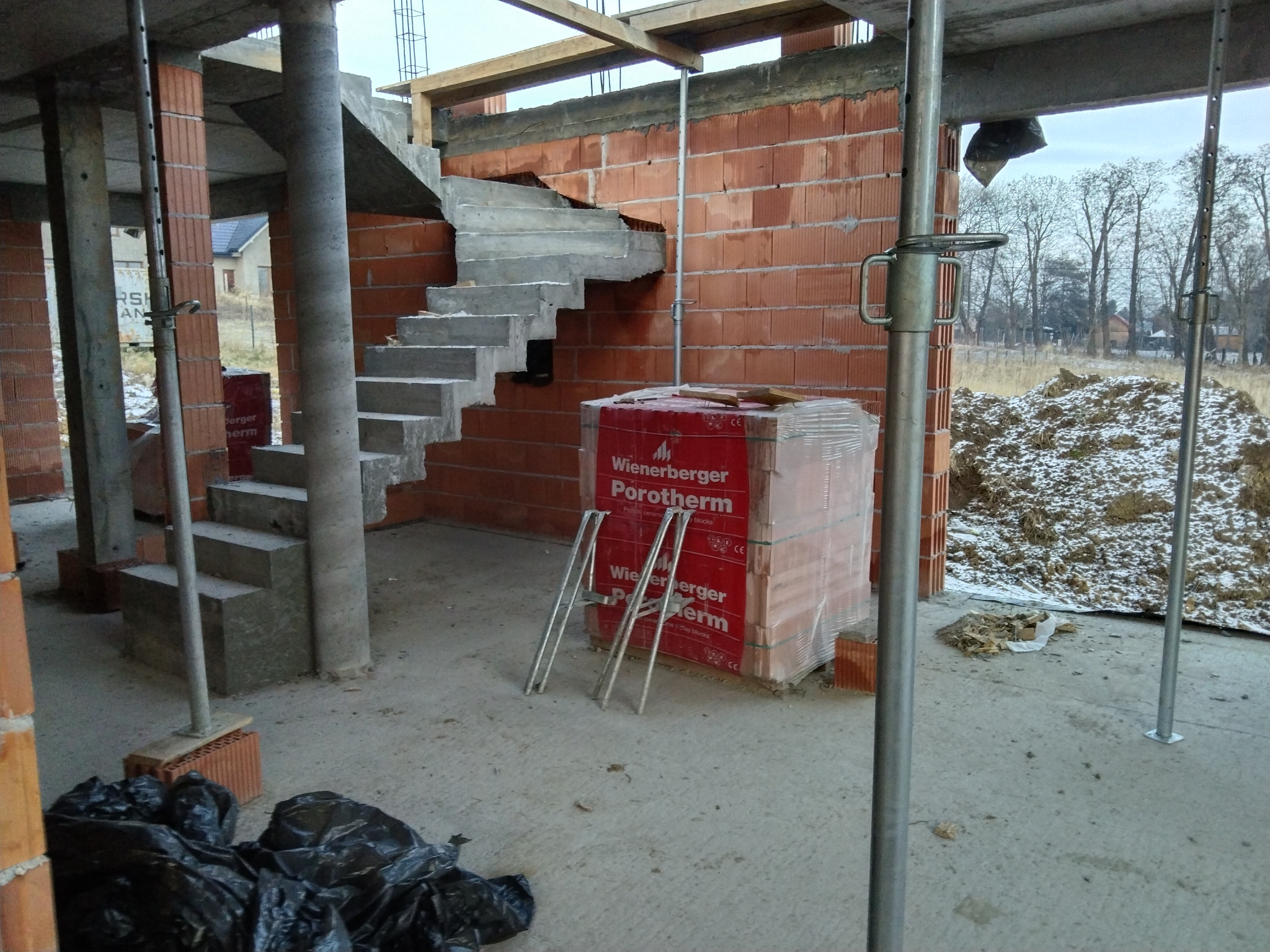 Surowy betonowy bieg schodów w trakcie budowy, widoczne czerwone cegły Porotherm, podpory stalowe, gruz i czarne worki na śmieci na surowym betonowym podłożu wewnątrz niedokończonego budynku.