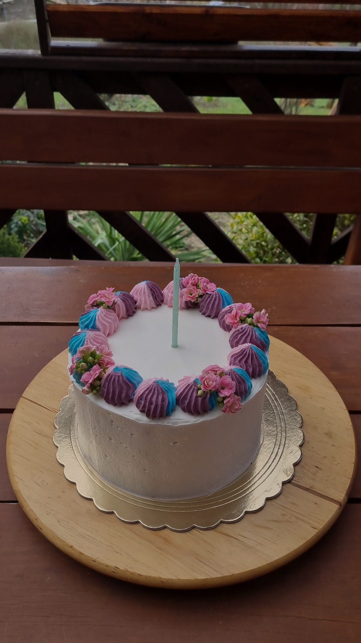 Tort z białym kremem, ozdobiony kolorowymi bezami (różowe, fioletowe, błękitne) i drobnymi różowymi kwiatkami, z pojedynczą zieloną świeczką, na złotej podkładce i drewnianym stole z widocznym...