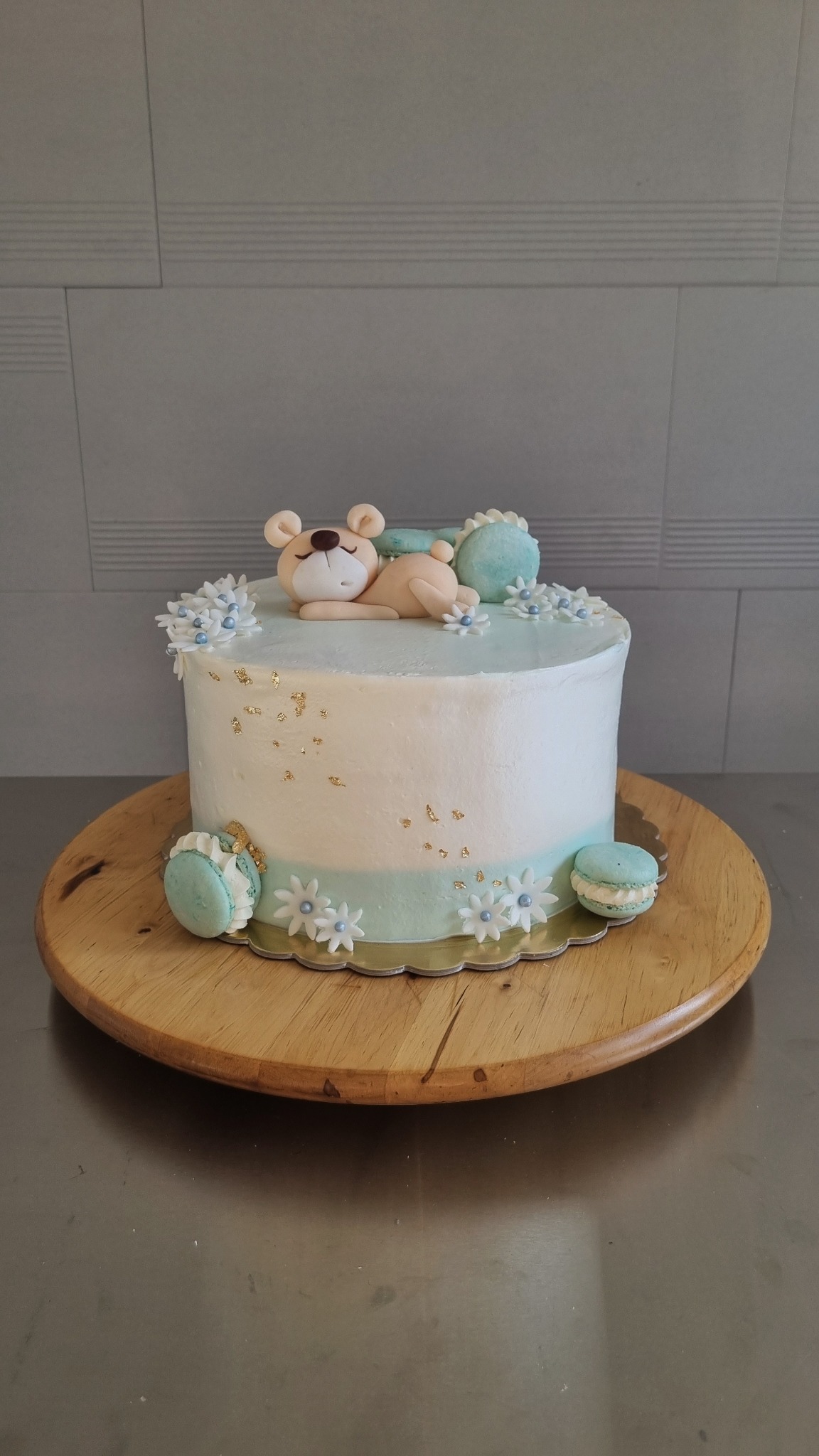 Tort z białym kremem, złotymi płatkami i śpiącym misiem z lukru, udekorowany małymi białymi kwiatkami i turkusowymi makaronikami, umieszczony na drewnianej tacy.
