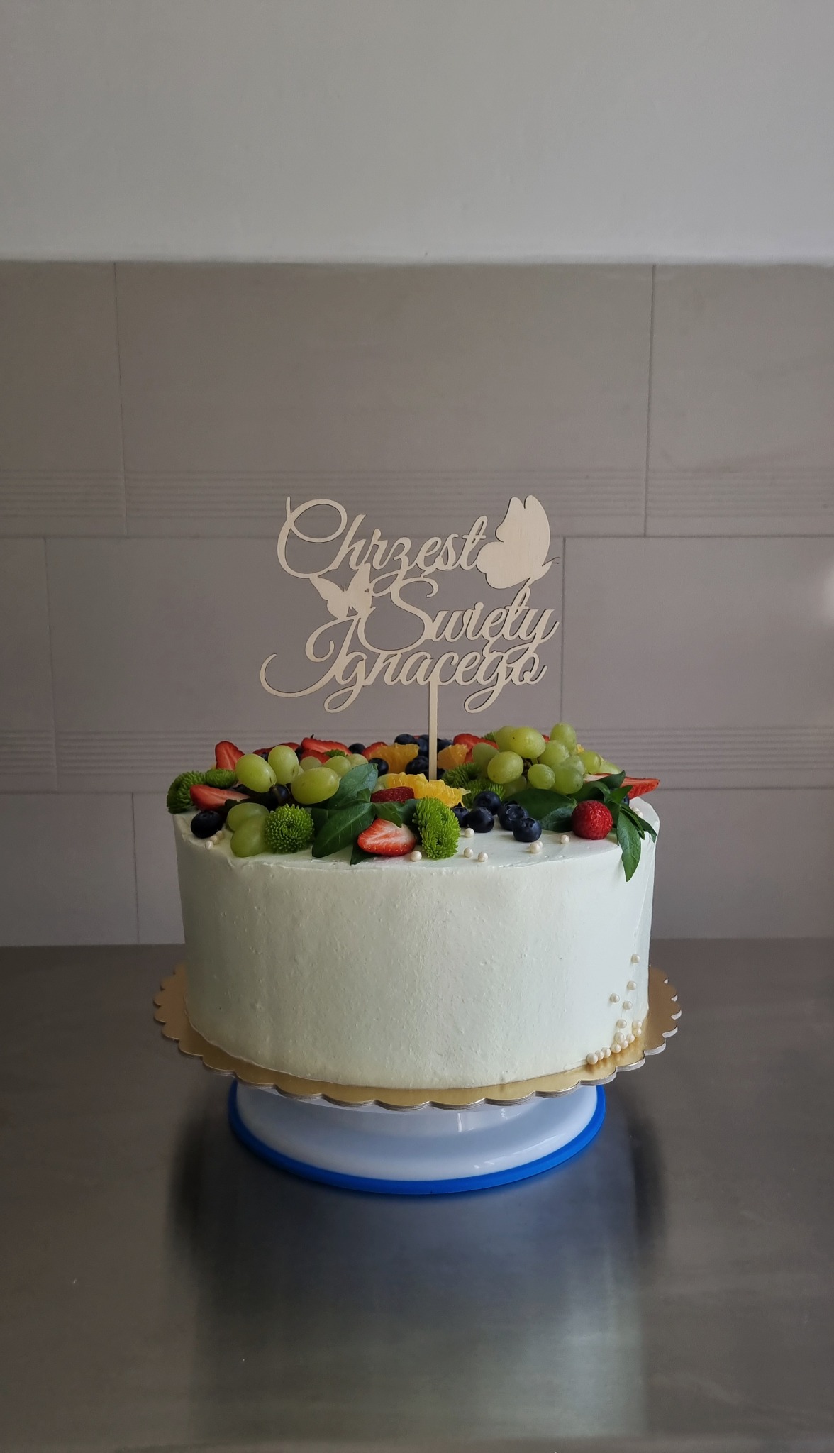 Tort z białym kremem, dekorowany świeżymi owocami i drewnianym topperem z napisem 'Chrzest Święty Ignacego', umieszczony na obrotowym stojaku.