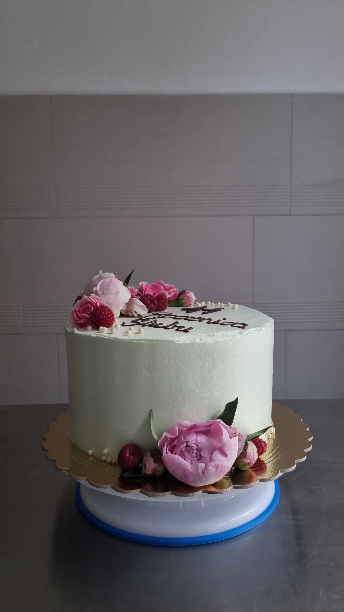 Tort z jasną polewą, udekorowany różowymi kwiatami, malinami i drobnymi perłami cukrowymi, z napisem wykonanym czekoladą, umieszczony na złotej paterze na obrotowej podstawie.