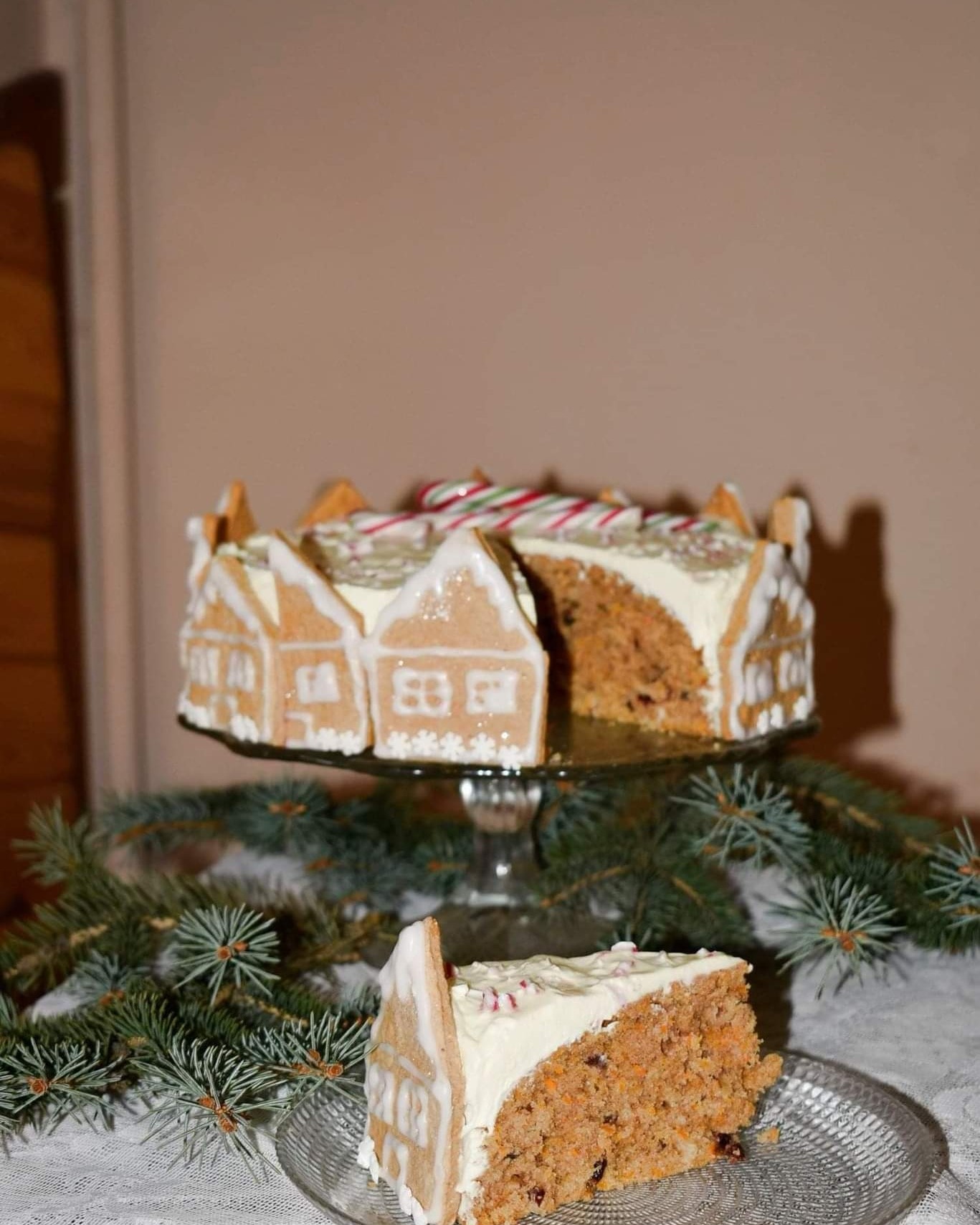 Torcik marchewkowy udekorowany piernikowymi domkami z białym lukrem, całość na szklanym stojaku, obok gałązki świerku i kawałek tortu na szklanym talerzyku