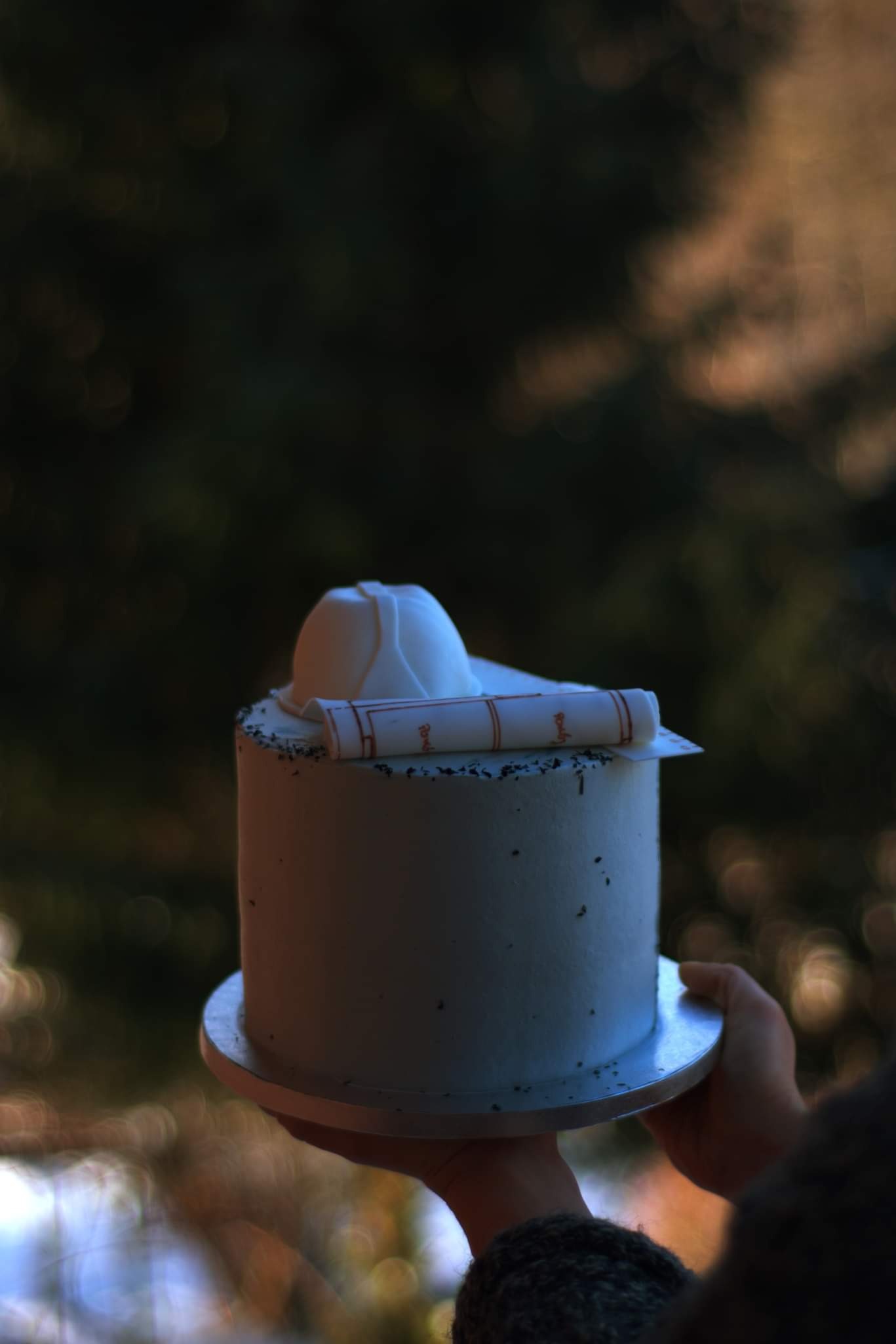 Tort z białym kremem, posypany ciemnymi kropkami, udekorowany jadalnym kaskiem budowlanym i zwiniętym planem architektonicznym, trzymany w dłoniach na tle rozmytego, zielonego tła.